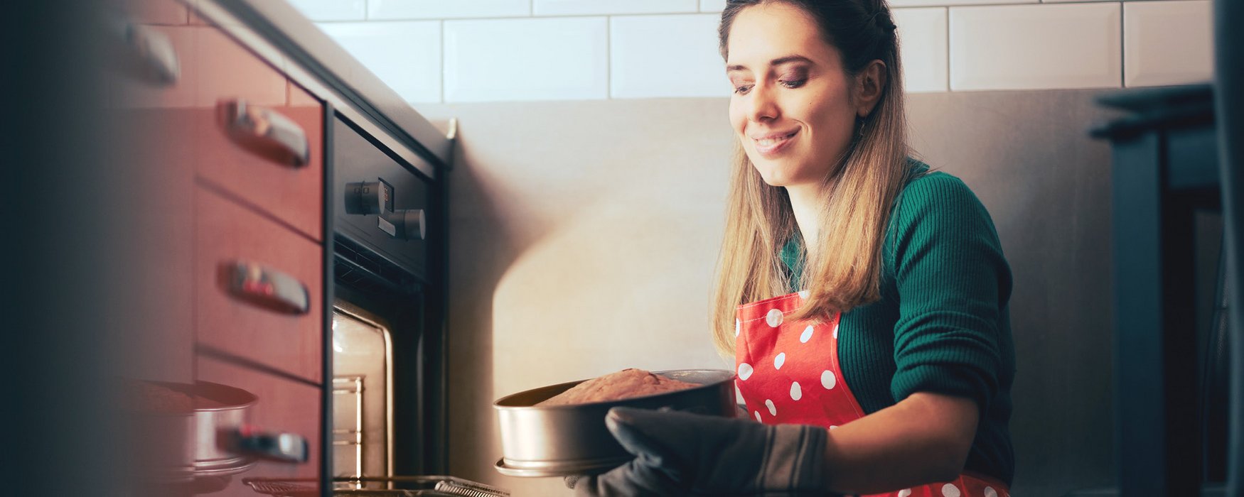 Junge Frau nimmt mit Backhandschuhen einen gebackenen Kuchen in einer grauen Form aus dem Ofen