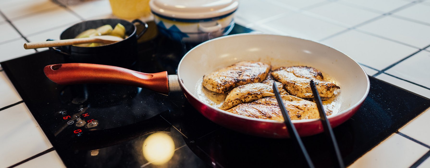 Pan, coated in red on the inside, white on the outside is standing on a stove with roasted meat chunks in it