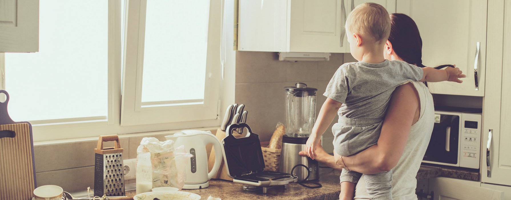 Mother with a toddler on her arm is standing in the kitchen and observes how the waffle iron is heating up 