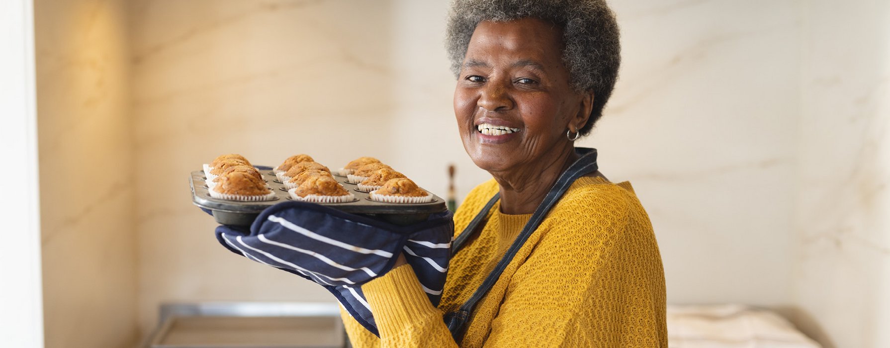 Elderly joyful woman is holding a muffin tray with baked muffins in it