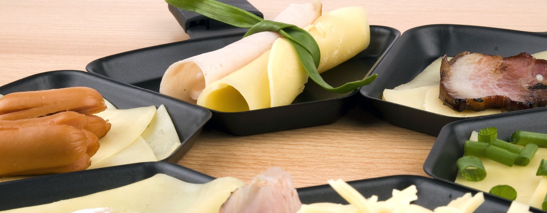 Schwarz beschichtete Raclette-Pfännen, die unterschiedlich befüllt sind, z.B. mit Käse, Wurst, Aufschnitt oder Gemüse