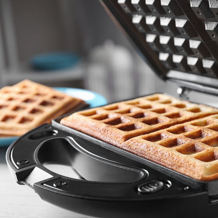 A baked waffle is laying in a black coated waffle iron and two baked waffles are on a blue plate next to it