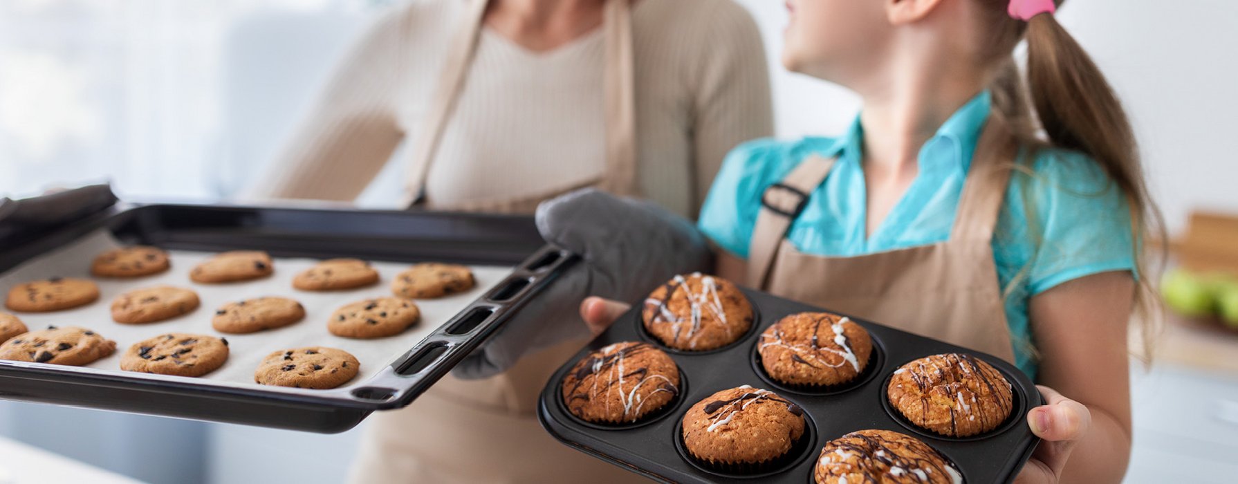 Frau und Mädchen präsentieren gemeinsam Gebackenes. Cookies auf einem Blech, Muffins in einer Backform