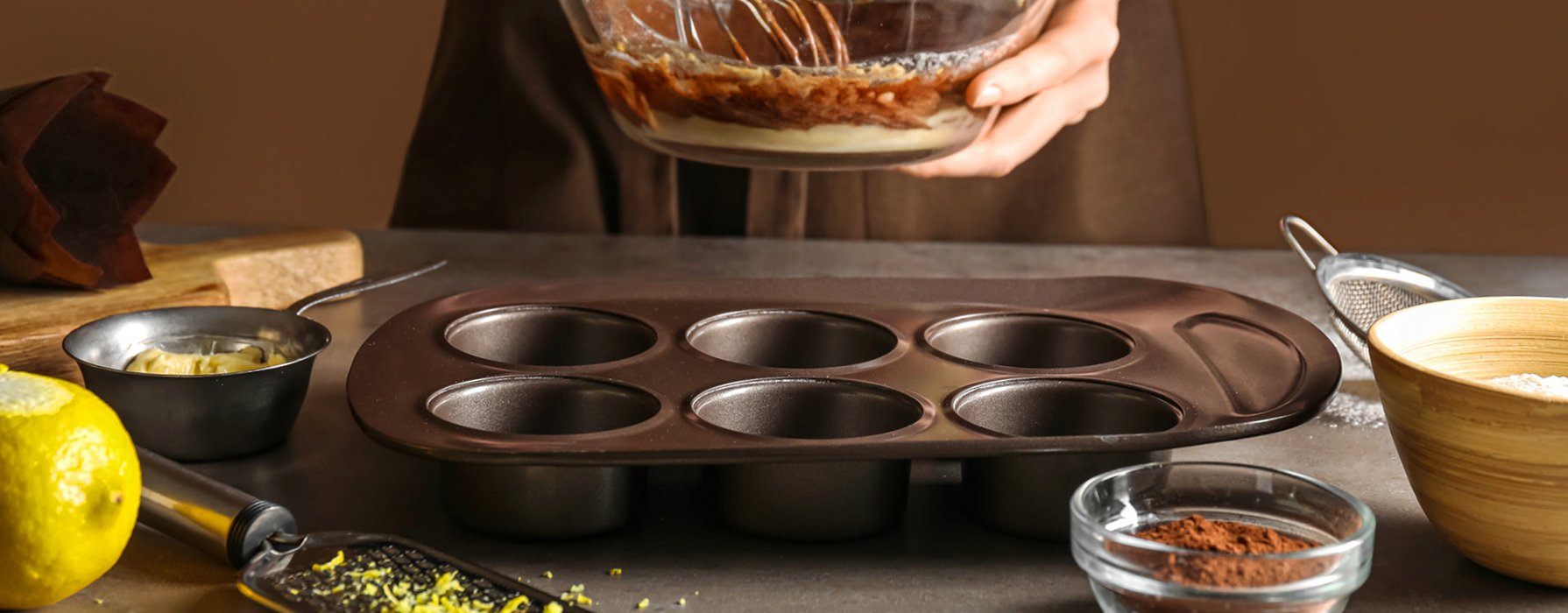 Auf dem Tisch ein schwarz beschichtetes Muffinformen-Blech, in das sogleich Schokoladenteig eingefüllt wird 