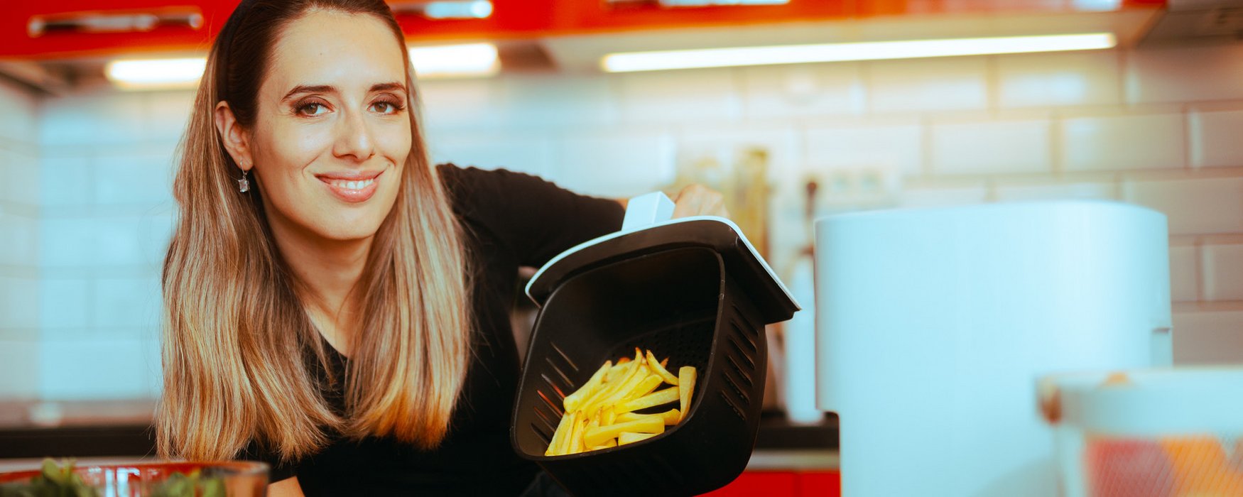 Junge Frau zeigt freudig die fertig gebackenen Pommes Frites in einem Korb der Heissluftfriteuse