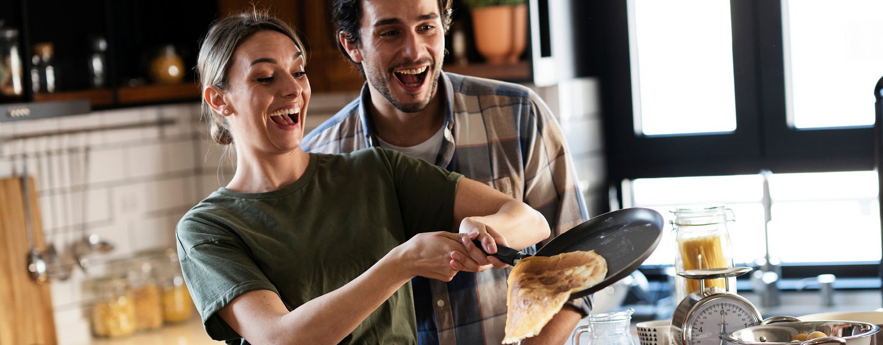 Junges lachendes Paar lässt einen Pfannkuchen aus einer schwarzen, beschichteten Pfanne gleiten