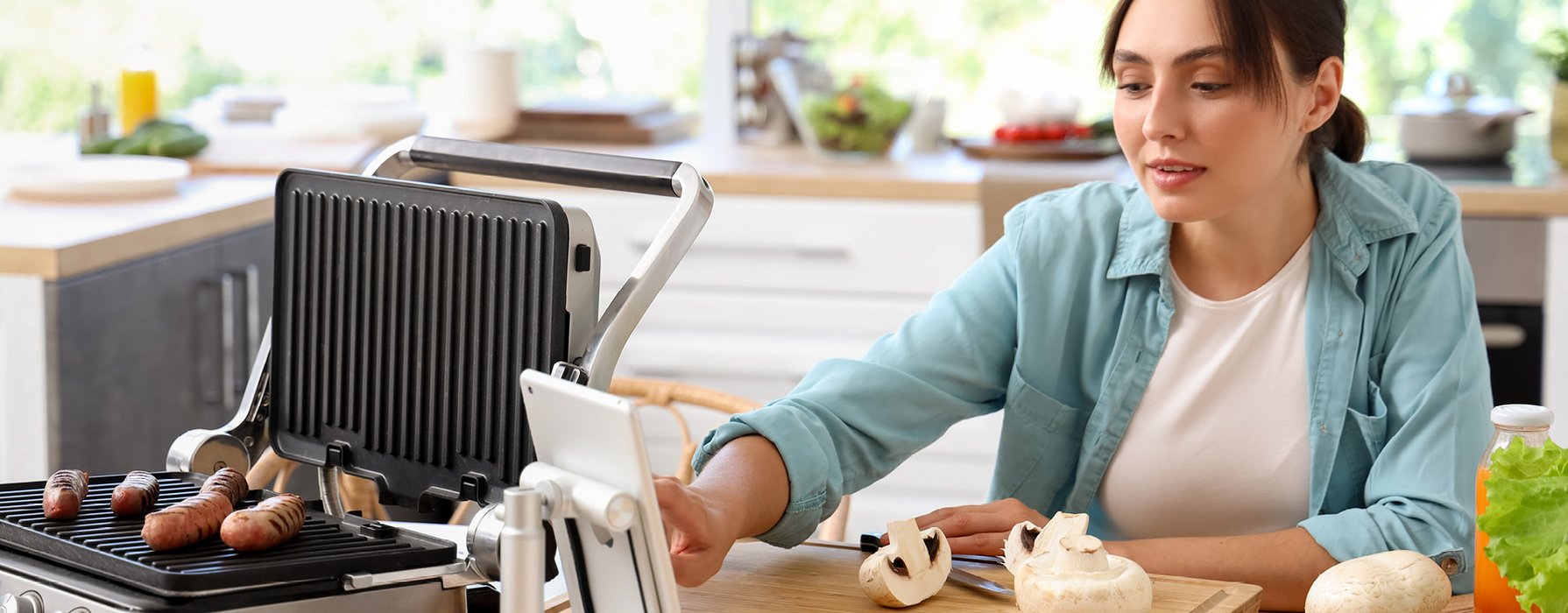 Woman is sitting on a table next to her there is a griddle with grilled sausages placed on the coated grill deflector plate