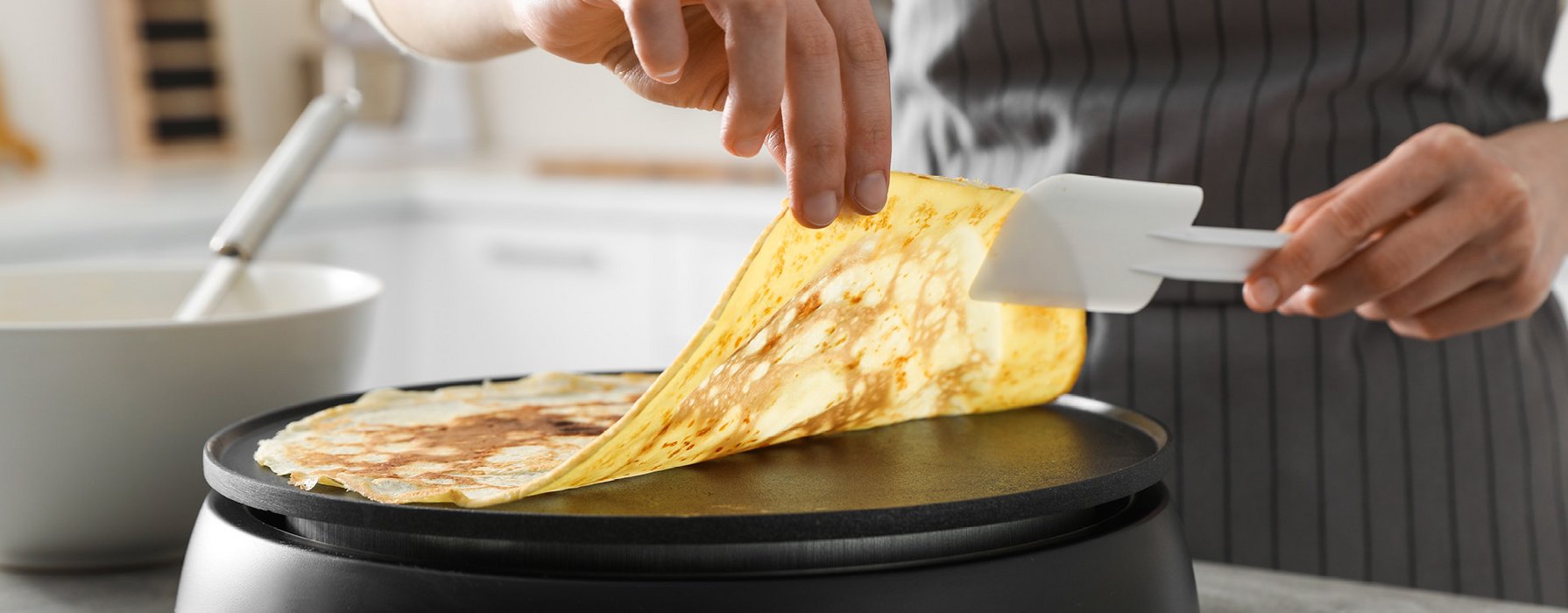 Eine gebratene Crêpe wird mit Hilfe eines Kunststoffschabers von der beschichteten Platte angehoben