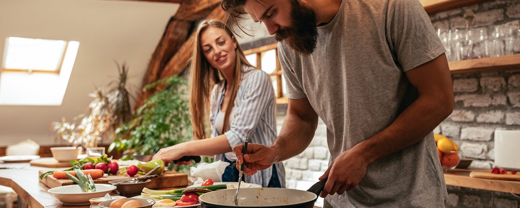 Young couple prepares food in a rustic kitchen. The woman cuts some vegetables, the man roasts meat in a frying pan.