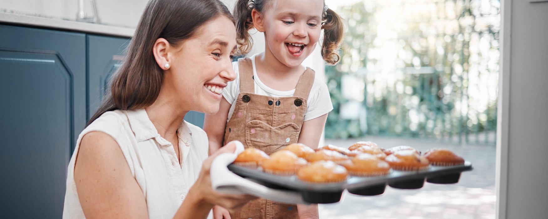 Junge Mutter nimmt gebackene Muffins in einer Form aus dem Ofen. Ein kleines Mädchen steht freudestrahlend daneben