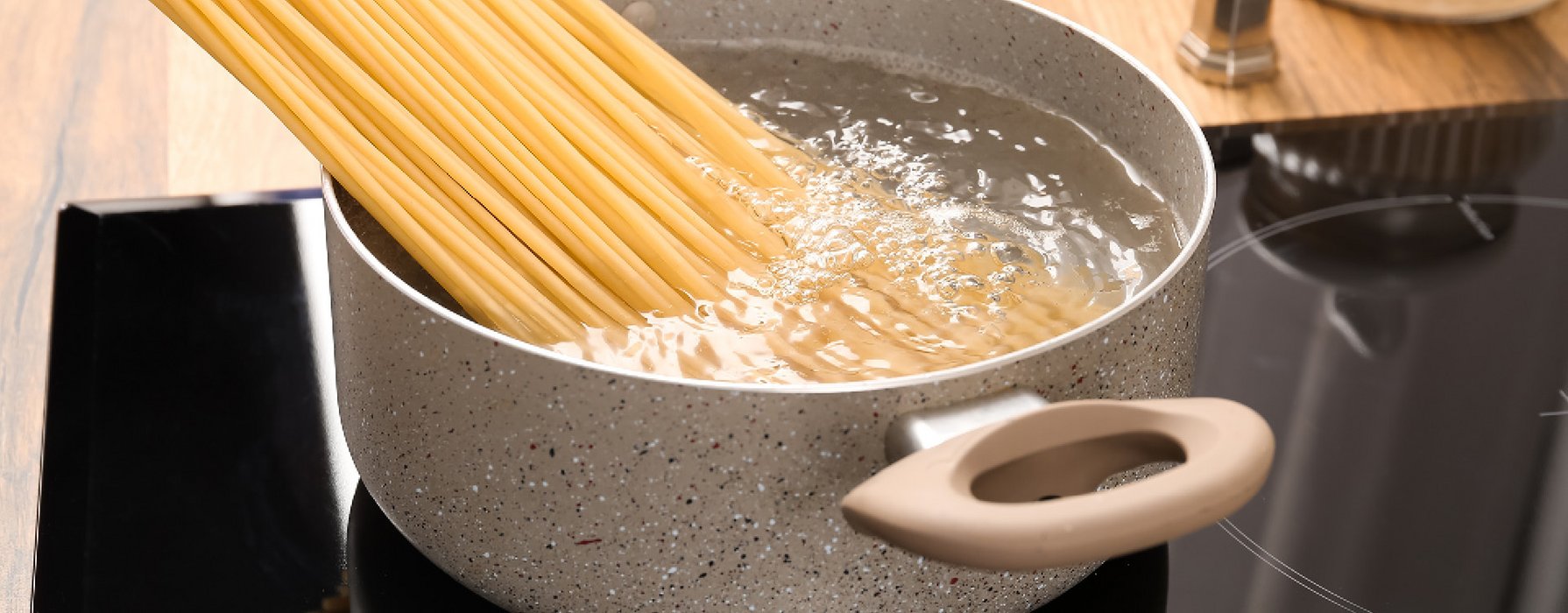 A pot with beige and black marbled coating is filled with spaghetti in boiling water and standing on the stove